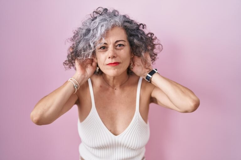 Middle age woman with grey hair standing over pink background trying to hear both hands on ear gesture, curious for gossip