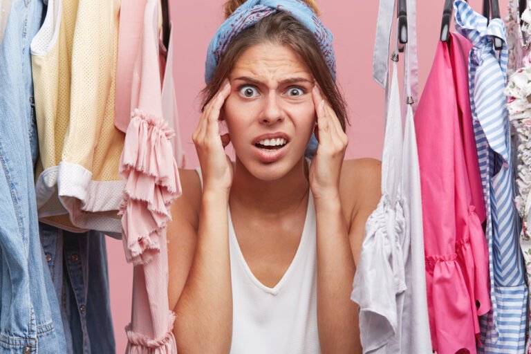 Nervous woman keeping hands on face, looking terrified while standing near wardrobe with clothes, realizing that she has nothing to wear for meeting with friends